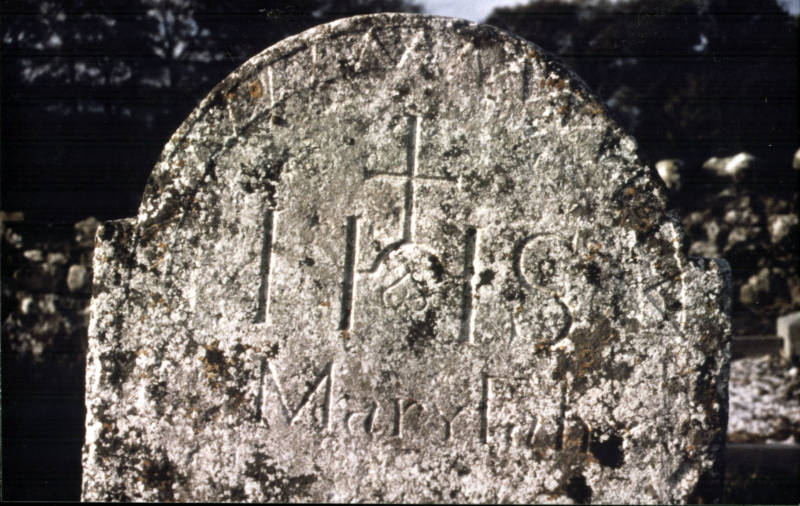 The omega memento mori symbol forming the cross-bar of the ‘H’ in the IHS monogram.